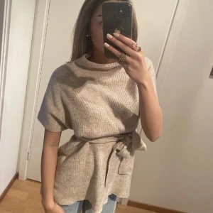 Stickad poncho med bälte - Säljer en stilren beige poncho med ribbad struktur och knytbälte i midjan. Perfekt för lager-på-lager under höst och vår. Den har en hög krage och är ärmlös, vilket gör den lätt att matcha med olika outfits. Pris kan diskuteras!!💘💘