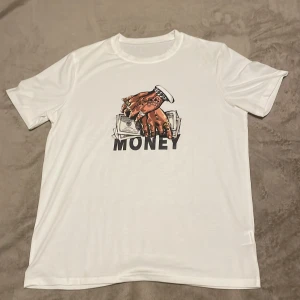 Vit t-shirt med tryck  - Säljer en vit t-shirt med ett coolt tryck av en hand som håller pengar och texten 'MONEY'. Perfekt för en avslappnad stil. T-shirten har korta ärmar och en rund halsringning.