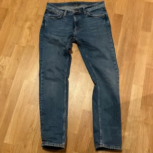 Blå jeans från Nudie Jeans (Lean Dean) - Snygga blå jeans från Nudie Jeans med klassisk femficksdesign och orange sömmar. De har en knappgylf och en bekväm passform som funkar till det mesta. Perfekta för dig som gillar stilrena och hållbara plagg.
