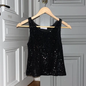 Svart paljettlinne - Säljer ett svart linne täckt med glittriga paljetter. Perfekt för en kväll ute eller en festlig tillställning. Linnet har en enkel design med breda axelband och ärmlös stil.