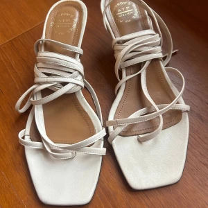 Vita sandaler med klack och snörning - Eleganta vita sandaler med fyrkantig tå och smal klack. Skorna har dekorativa snören som kan knytas runt vristen för en stilfull look. Perfekta för sommarens festligheter.