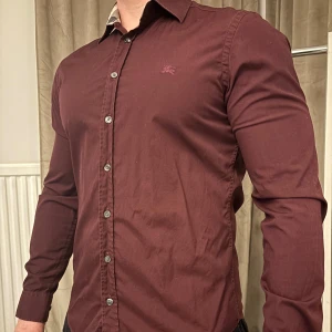 Vinröd skjorta från Burberry Brit - Snygg vinröd skjorta från Burberry Brit med broderad logga på bröstet. Skjortan har långa ärmar och knappar framtill. Kragen har ett klassiskt rutigt mönster på insidan som ger en stilren touch.