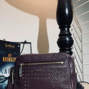 Plommonförgaf axelväska från DKNY - Jättesnygg plommonförgaf väska från DKNY med dragkedja och nitar på framsidan. Perfekt för att ge din outfit en uppgradering. Väskan har en justerbar axelrem och är tillverkad i ett strukturerat material.