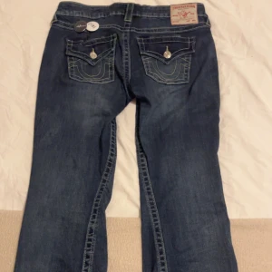 Blå jeans från True Religion - Snygga blå jeans från True Religion med klassisk femficksdesign och dekorativa sömmar på bakfickorna. Jeansen har en knappgylf och är i en stilren bootcut-modell. Perfekta för en avslappnad look.  Modellen är joey! 