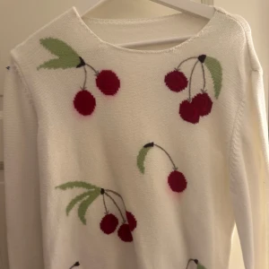 Vit tröja med körsbärsmönster - Charmig vit tröja med röda körsbär och gröna blad som mönster. Tröjan har långa ärmar och en rund halsringning. Perfekt för en söt och lekfull stil. Hemstickad.