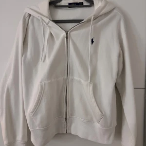 Vit kofta från Ralph Lauren - Säljer en stilren vit kofta från Ralph Lauren med dragkedja och klassisk logga på bröstet. Tröjan har en bekväm passform med långa ärmar och en praktisk huva. 