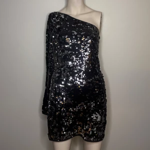 Svart paljettklänning från Jones + Jones - Svart one-shoulder klänning från Jones + Jones, täckt med glittriga paljetter. Klänningen är kort och har en lång ärm på ena sidan. Passar en storlek S.
