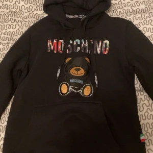 Svart hoodie från Moschino med björndetalj - Säljer en svart hoodie från Moschino med en söt björndetalj på framsidan. Hoodien har en färgglad Moschino-logga och en praktisk känguruficka. Perfekt för en avslappnad stil.
