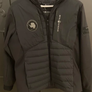 Svart jacka från Sail Racing - Snygg svart jacka från Sail Racing med coola patchar på bröstet. Jackan har en quiltad design och dragkedja framtill. Perfekt för kyligare dagar med sin långa ärmar och huva.