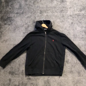 Svart hoodie från Polo Ralph Lauren - Säljer en stilren svart hoodie från Polo Ralph Lauren med dragkedja och det klassiska röda logotypen på bröstet. Perfekt för en avslappnad stil. Den har långa ärmar och en bekväm huva.