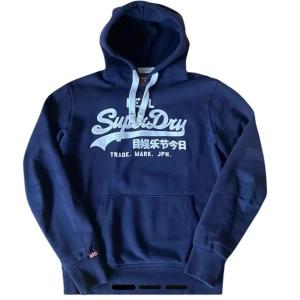 Blå hoodie från Superdry - Säljer en snygg blå hoodie från Superdry med vit text på framsidan. Den har en klassisk känguruficka och justerbar huva med snören. Perfekt för en avslappnad stil.