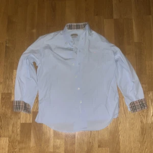 skjorta från Burberry - Snygg skjorta från Burberry med klassiskt rutigt mönster på insidan av kragen och ärmsluten. Skjortan har en broderad logga på bröstet och knappar framtill. Perfekt för en stilren look.