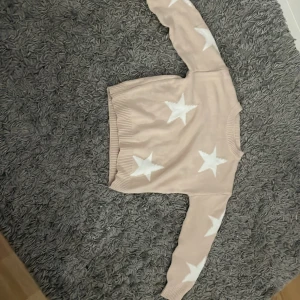 Beige stickad tröja med stjärnor - Säljer en mysig beige stickad tröja med vita stjärnor. Tröjan har långa ärmar och ribbade muddar vid ärmslut och nederkant. Perfekt för kyliga dagar!