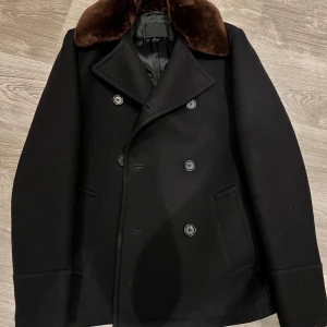 Ull Caban Kappa Prada Nypris 52k! - cabanjacka i ull med pragmatisk elegans definieras av eleganta linjer och detaljer som uttrycker en raffinerad, modern attityd. Med avtagbar pälskrage.   Mycket fint skick endast använd ett fåtal gånger och är så gott som ny!  Nypris cirka 52000kr!  Mitt pris: 17500kr Tillbehör: Lappar     En Cabanjacka: Tjockt ulltyg – Vanligtvis gjord av kraftig ull för att ge värme och skydd mot vind och kyla. Bred krage – Kan fällas upp för extra skydd mot väder och vind. Kort till medellång längd – Slutar o