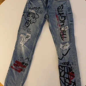 Jeans med graffiti-design från Bershka - Unika jeans från Bershka med graffiti-inspirerad design i svart och rött. De har en straight passform och är tillverkade i denim. Byxorna har slitna detaljer och tryck med text och mönster som ger en edgy look.