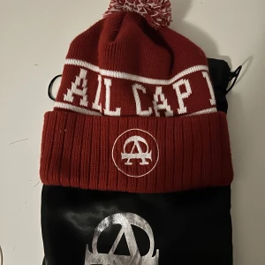 Röd stickad mössa med tofs - Snygg röd stickad mössa med vit text och logotyp framtill. Mössan har en vit rand och en matchande tofs på toppen. Perfekt för kalla dagar.