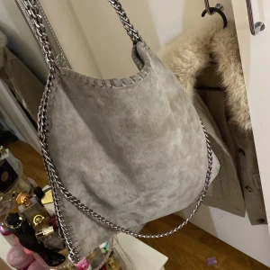 Grå axelväska med kedjedetaljer - Snygg grå axelväska med kedjedetaljer längs kanterna och axelremmen. Väskan har en dragkedja på baksidan. Väldigt bra skick använd endast 2-3 gånger 