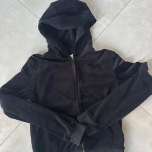 Svart hoodie från Juicy Couture - Snygg svart hoodie från Juicy Couture med dragkedja och huva. Perfekt för en avslappnad stil. Den är tillverkad i ett mjukt material som ger en bekväm känsla. 12- 13 år,storlek