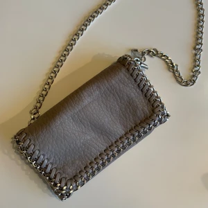 Tiamo väska  - Grå väska från tiamo med axelbands kedja. Väskan har två innerfack varav ett är med dragkedja. Liten modell. 