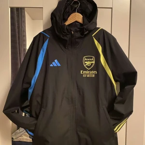 Svart träningsjacka från Adidas - Säljer en svart träningsjacka från Adidas med Arsenal-logga och Emirates-tryck. Jackan har blå och gula detaljer samt en dragkedja framtill. Perfekt för fotbollsfans! 💙💛