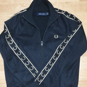 Svart track jacket från Fred Perry - Nypris - 1800 KR                         Sitter som XS. Snygg svart track jacket från Fred Perry med dragkedja och ikoniska lagerkransar längs ärmarna. Perfekt för en sportig look med stil. Jackan har en normal passform och är tillverkad i ett mjukt syntetmaterial.
