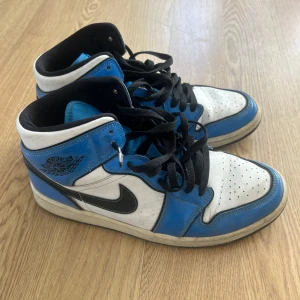 Nike Air Jordans 1 - Snygga Nike Air Jordans i blått och vitt med svart snörning. Skorna har en hög modell med perforerade detaljer på tån och en ikonisk Nike-logga på sidan. Perfekta för dig som vill ha en stilren och sportig look. Ganska använda storlek 43