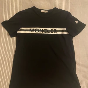 Svart t-shirt från Moncler - Snygg svart t-shirt från Moncler med deras logga tryckt i vitt över bröstet och en broderad logga på ärmen. Perfekt för en stilren look.