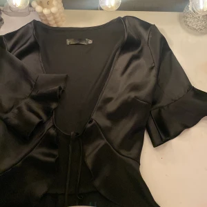 Svart satinbolero med volangdetaljer - Snygg svart bolero i satin med volangdetaljer och knytning framtill. Den har korta ärmar och en v-ringad design som ger en elegant look. Perfekt för att lyfta vilken outfit som helst. Jättefint med t.ex ett linne under! Inga defekter