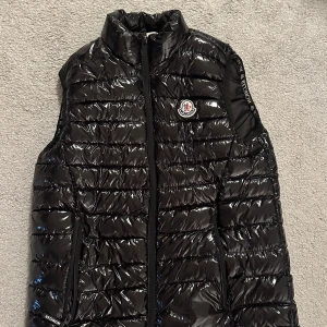 Svart dunväst från Moncler - Snygg svart dunväst från Moncler med glansig yta och dragkedja framtill. Västen har Moncler-logga på bröstet och är perfekt för kyligare dagar. Kom med bud