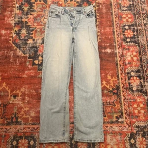 Levi's Ribcage Straight Ankle Jeans - Snygga ljusblå Levi's Ribcage Straight Ankle jeans med hög midja och knappgylf. Det är rivna där bak. Perfekta för en avslappnad stil med en rak passform som slutar vid ankeln. Klassisk Levi's-design med ikonisk läderpatch baktill.