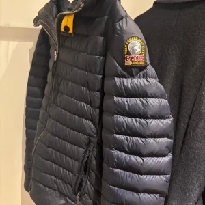 Svart pufferjacka från Parajumpers - Säljer en snygg blå pufferjacka från Parajumpers med dragkedja och en cool patch på ärmen. Jackan har en quiltad design och är perfekt för kyligare dagar. Den har en praktisk dragkedja framtill och en unik stil med detaljer i gult och orange.