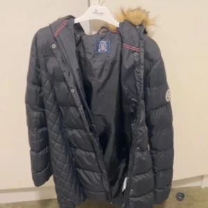 Svart quiltad jacka med pälskrage - Snygg svart quiltad jacka med pälskrage och huva. Jackan har en dragkedja framtill och är perfekt för kyligare väder. Den har en klassisk design med långa ärmar och en bekväm passform.