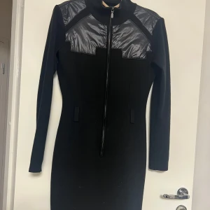 Svart klänning med dragkedja - Snygg svart klänning med dragkedja framtill och glansiga detaljer på axlarna. Den har en tight passform och långa ärmar, perfekt för en stilren look. Klänningen är både elegant och modern.