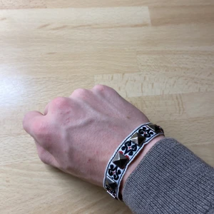 Svart och vitt armband med nitar - Unikt armband med svart och vitt mönster och dekorativa nitar. Armbandet har justerbara snören för perfekt passform och en röd detalj som ger en extra touch. Perfekt för att ge din outfit en edgy stil.