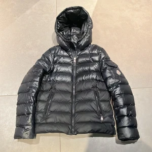 Svart dunjacka från Moncler - Säljer en snygg svart dunjacka från Moncler med huva och dragkedja. Jackan har ett broderat Moncler-märke på ärmen och praktiska fickor med dragkedja. Perfekt för kyliga dagar!