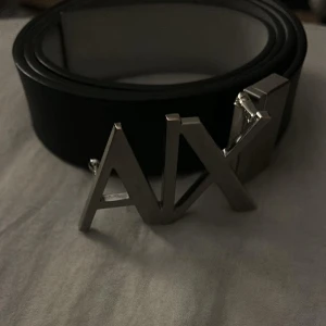Svart/vit armani bälte - Snyggt svart bälte från Armani Exchange med en iögonfallande silverfärgad spänne i form av bokstäverna 'AX'. Perfekt för att ge en stilren touch till din outfit.