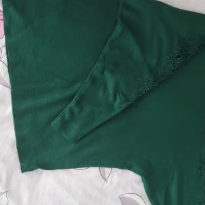 Grön tröja med spetsdetaljer från AnFaiya - Säljer en elegant cashmere blandat grön tröja från AnFaiya med vackra spetsdetaljer längs ärmarna. Tröjan har en normal passform och är perfekt för att ge en stilfull touch till din garderob. Passar bra till både vardag och speciella tillfällen.