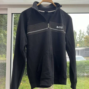 Svart hoodie från BOSS - Snygg svart hoodie från BOSS med dragkedja och prickigt mönster över bröstet. Perfekt för en stilren och avslappnad look. Den har långa ärmar och praktiska fickor framtill.