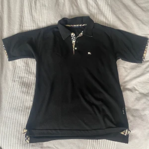 Svart pikétröja från Burberry - Säljer en stilren svart pikétröja från Burberry med klassiskt rutigt mönster på insidan av kragen och knappslån. Tröjan har korta ärmar och en liten broderad logotyp på bröstet. Perfekt för en elegant och avslappnad look.