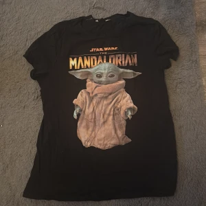 Svart Star Wars The Mandalorian t-shirt - Säljer en svart t-shirt med tryck från Star Wars: The Mandalorian. T-shirten har en bild av Baby Yoda på framsidan och texten 'The Mandalorian' i orange och gult. Perfekt för fans av serien!