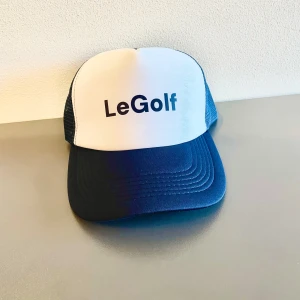 Vit och blå golfkeps från LeGolf UF - Snygg keps från LeGolf med vit framsida och blått nätmaterial på baksidan. Kepsen har en justerbar rem för perfekt passform och texten 'LeGolf' tryckt på framsidan. Perfekt för golfentusiaster men också vardags användning!
