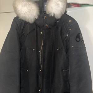 Svart dunjacka från Moose Knuckles - Säljer en svart dunjacka från Moose Knuckles med en lyxig pälsdetalj på huvan. Jackan har en dragkedja och knappar framtill samt ett coolt emblem på ärmen. Perfekt för kalla vinterdagar. STL M 