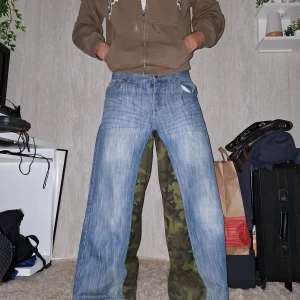 Blå jeans med camo-detaljer från S&J Denim - W34 L32 Unika blå jeans från S&J Denim med coola camo-detaljer på bakfickorna. Byxorna har en loose passform och är perfekta för en avslappnad stil. De har en knappgylf och tydliga sömmar som ger en extra touch.