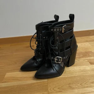 Prettylittlething svarta boots med klack och snörning - Snygga svarta boots i skinn med klack och spetsig tå. De har snörning och spännen som ger en edgy look. Perfekta för att lyfta vilken outfit som helst.