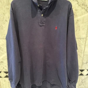 Mörkblå pikétröja från Ralph Lauren - Säljer en klassisk mörkblå pikétröja från Ralph Lauren i bomull. Tröjan har långa ärmar och en broderad röd logotyp på bröstet. Perfekt för en stilren look.