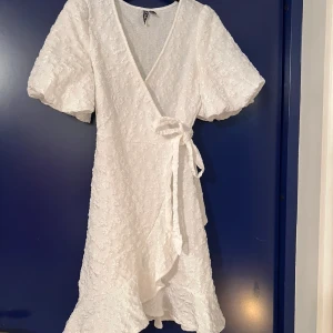 Vit omlottklänning från Pieces - Säljer en söt vit omlottklänning från Pieces med korta puffärmar och volangdetaljer. Klänningen har en knytning i midjan och ett diskret blommönster som ger en romantisk touch. Perfekt för sommarens alla tillfällen! 🌸Jag är 167