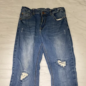 Blå jeans med slitningar från LAB Industries - Säljer ett par blå jeans från LAB Industries med coola slitningar på framsidan. De har en klassisk femficksdesign och knappgylf. Det finns även resår i midjan så man kan justera storleken lite.