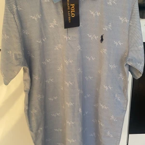 Blå pikétröja från Ralph Lauren - Säljer en stilren blå pikétröja från Ralph Lauren med ett subtilt vitt mönster och den klassiska loggan broderad på bröstet. Perfekt för en avslappnad men ändå elegant look. Tröjan har korta ärmar och en klassisk krage.