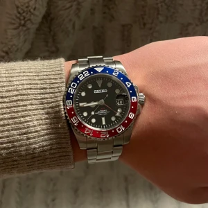 Pepsi Seiko Mod - Säljer en tvär snygg Seiko klocka med en iögonfallande blå och röd bezel. Som efterliknar ”Pepsi” Klockan har en svart urtavla med datumvisning och en robust metallarmband. 40mm. Perfekt för den som vill ha en stilren och funktionell klocka. Klockan är automatisk, med en NH34 movement. Alla länkar tillkommer såklart vid köp. 