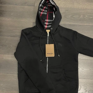 Svart huvtröja med dragkedja från Burberry - Snygg svart huvtröja från Burberry med dragkedja och klassiskt rutigt mönster på insidan av huvan. Tröjan har långa ärmar och praktiska fickor framtill. Perfekt för en stilren look.